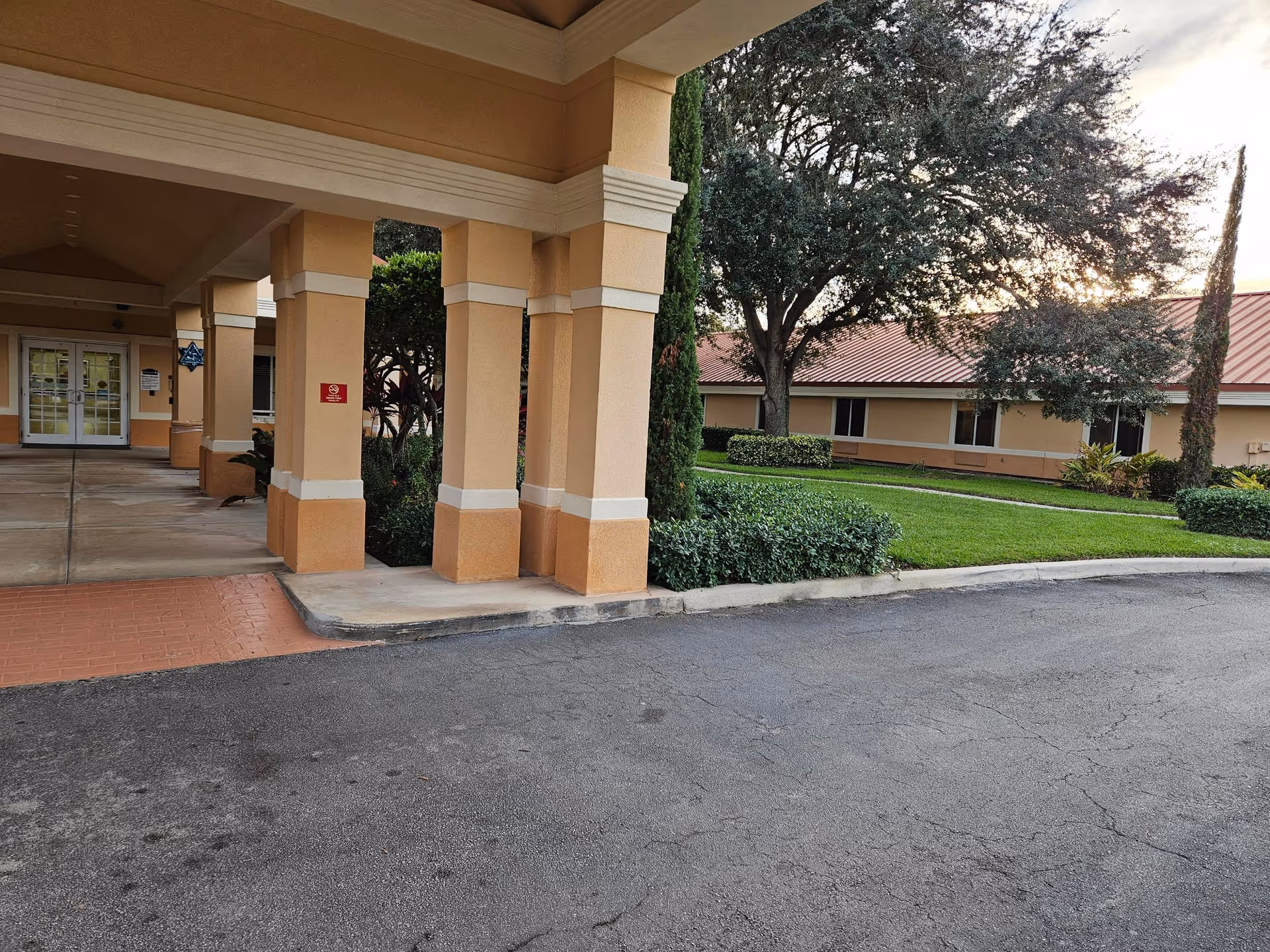 Covered porte-cochère and driveway at the front of a beige rehabilitation facility with columns, lawn, and trees.