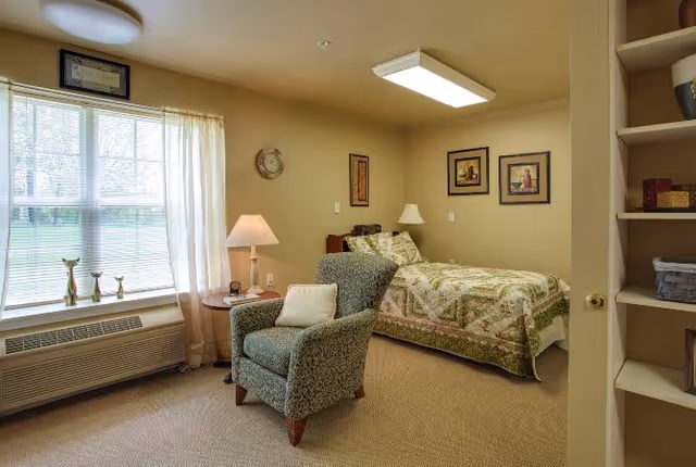 A cozy bedroom in a senior living facility with a single bed covered in a patterned quilt, a comfortable upholstered armchair with a pillow, a small round side table with a lamp, and a large window with sheer curtains letting in natural light. The walls are decorated with framed artwork and a clock, and there is a built-in shelving unit partially visible on the right.