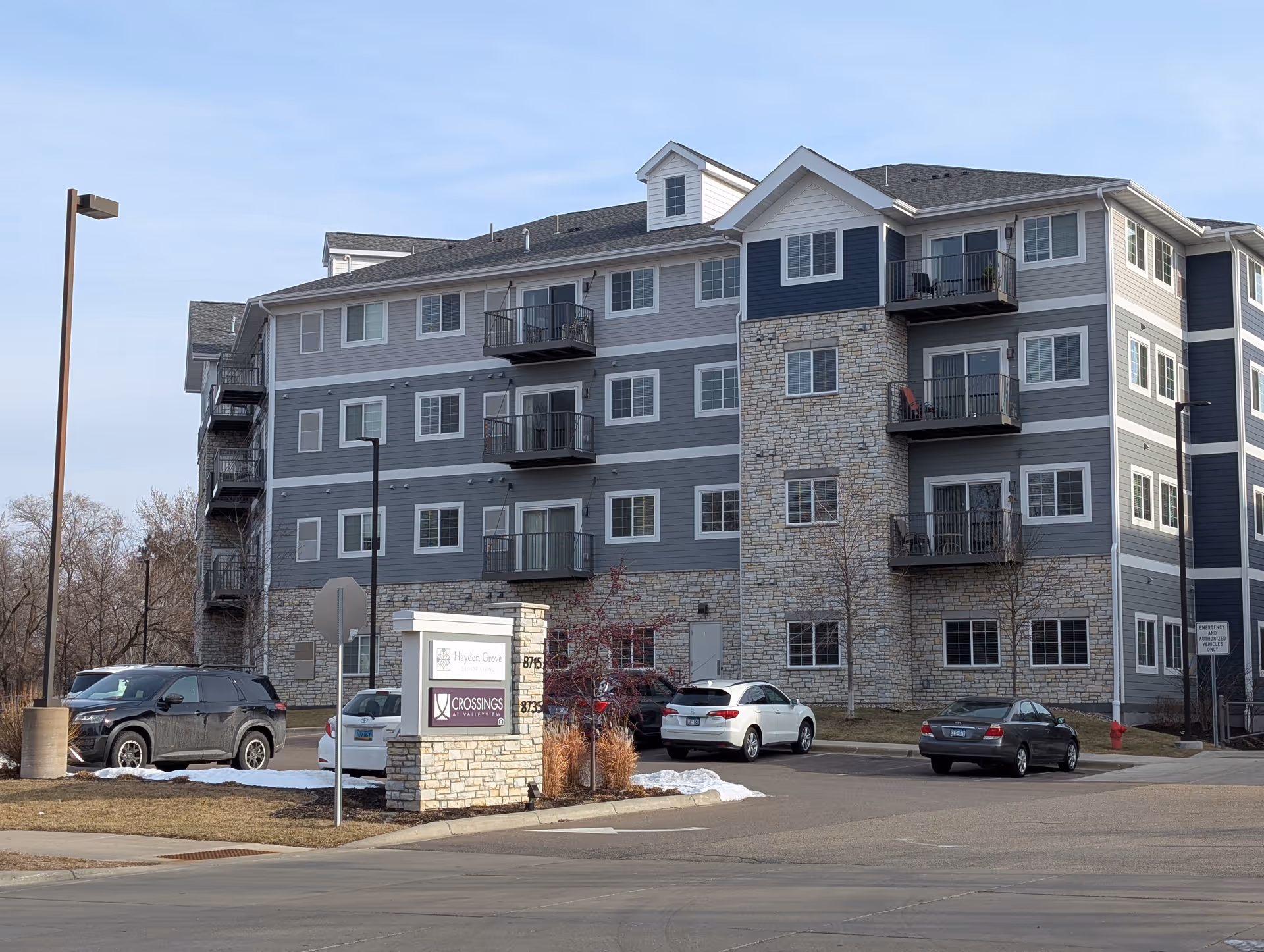 Exterior view of a multi-story senior housing building named Hayden Grove Senior Living with stone and gray siding, balconies, parked cars, and a sign at the entrance.