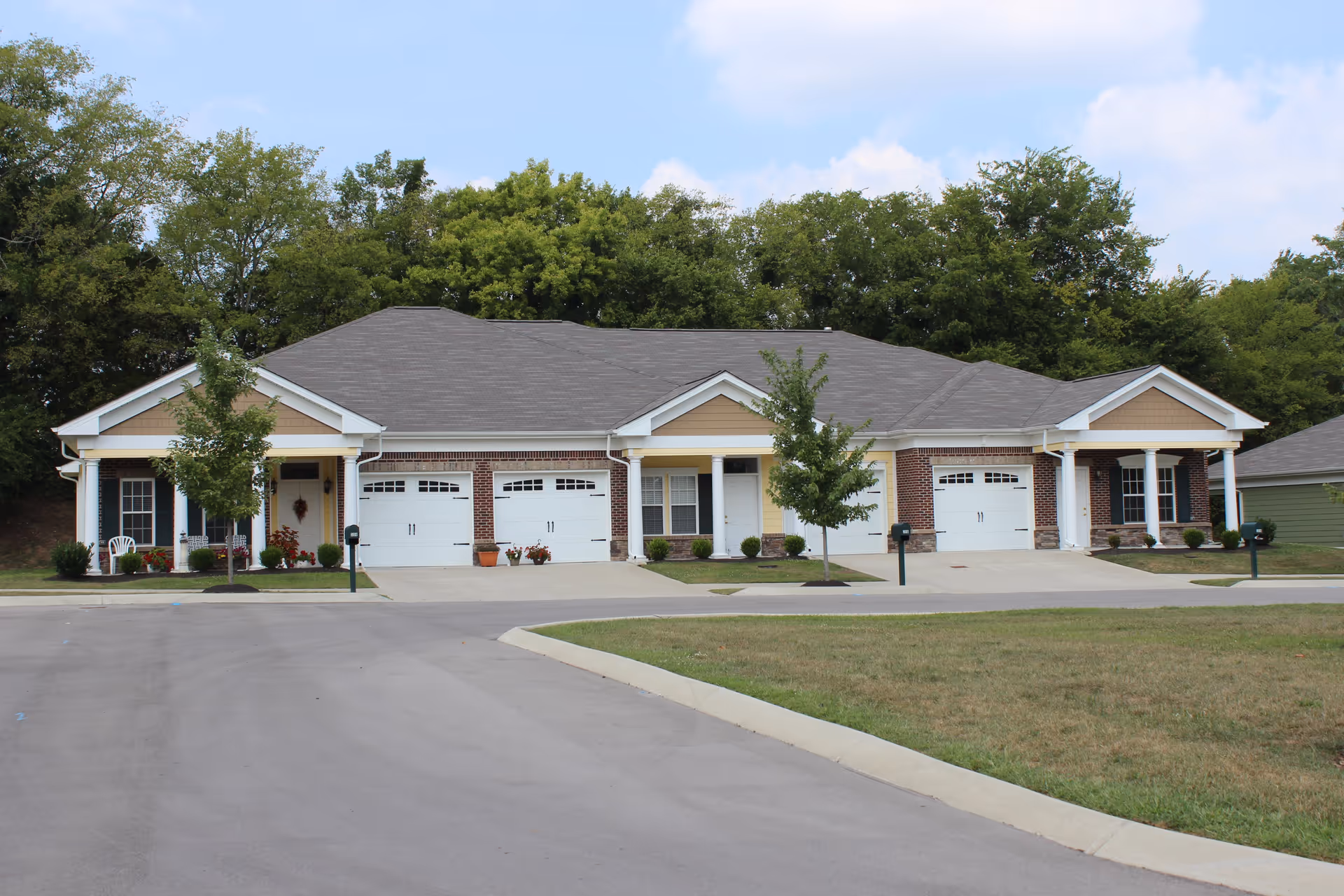 Exterior view of a single-story senior living facility building with three attached units, each featuring a garage with white doors, small front porches with columns, and landscaped areas with small trees and shrubs. The building has a gray shingled roof and is set against a backdrop of green trees under a partly cloudy sky.