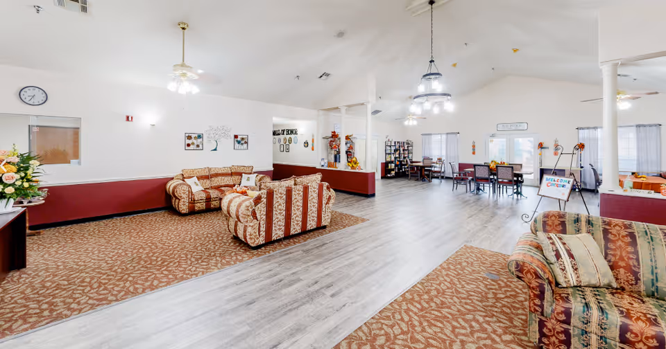 A spacious and well-lit common area in an assisted living facility featuring patterned sofas arranged on carpeted sections, wooden flooring, multiple tables and chairs, decorative wall art, ceiling fans with lights, and large windows with sheer curtains allowing natural light.
