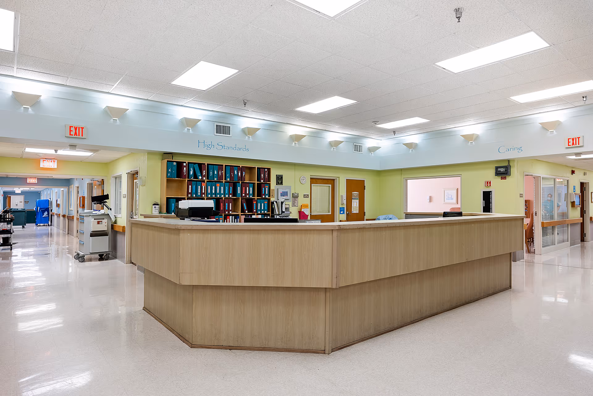 A clean and well-lit reception desk area in a senior living facility with shelves of organized binders behind the desk. The walls are painted light green and white, with exit signs visible above doorways. The ceiling has multiple fluorescent lights, and the floor is shiny and reflective.