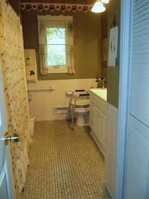 Narrow accessible bathroom with toilet, sink, grab bars, tiled floor, and window.