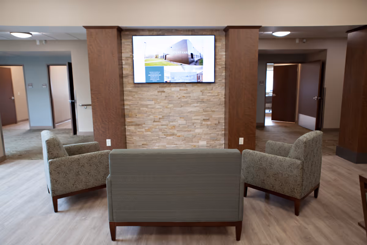 A cozy seating area in a senior living facility with a loveseat and two armchairs arranged around a wall-mounted flat screen TV. The TV is mounted on a stone accent wall framed by wooden panels. The room has light wood flooring and open doorways leading to other rooms in the background.