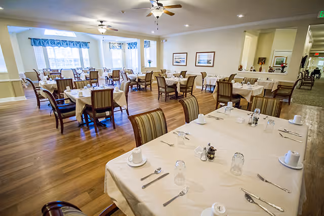 A spacious dining room with multiple tables covered in white tablecloths, each set with cups, glasses, and silverware. The room has wooden flooring, large windows with blue valances allowing natural light, ceiling fans, and framed artwork on the walls.