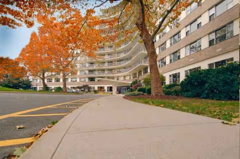 Exterior view of a curved senior living building with a walkway and autumn trees.