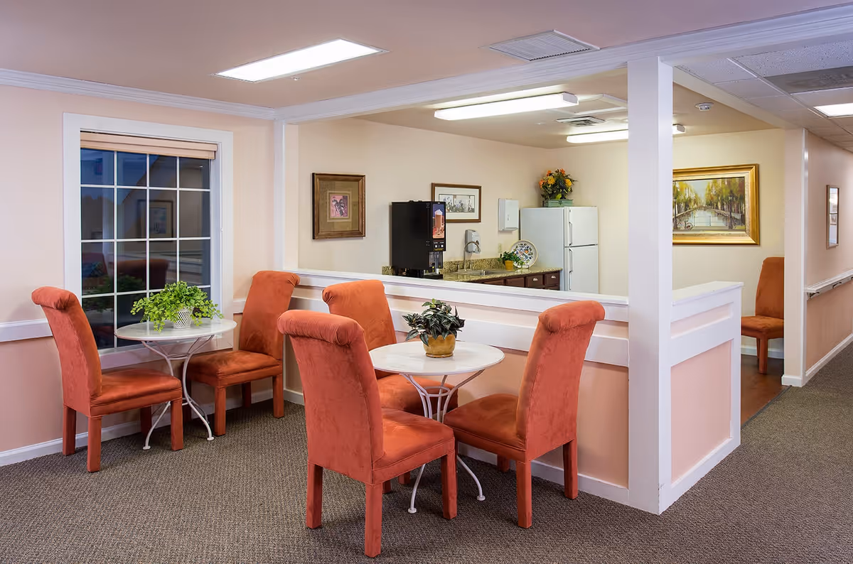 A cozy seating area with two small round tables, each surrounded by four orange upholstered chairs. Each table has a small potted plant on it. Behind the seating area is a half wall with a large window opening into a kitchen area that contains a refrigerator, a coffee machine, and some decorative items. The walls are light peach with white trim, and there are framed paintings hanging on the walls. The floor is carpeted, and the space is well-lit with ceiling lights.