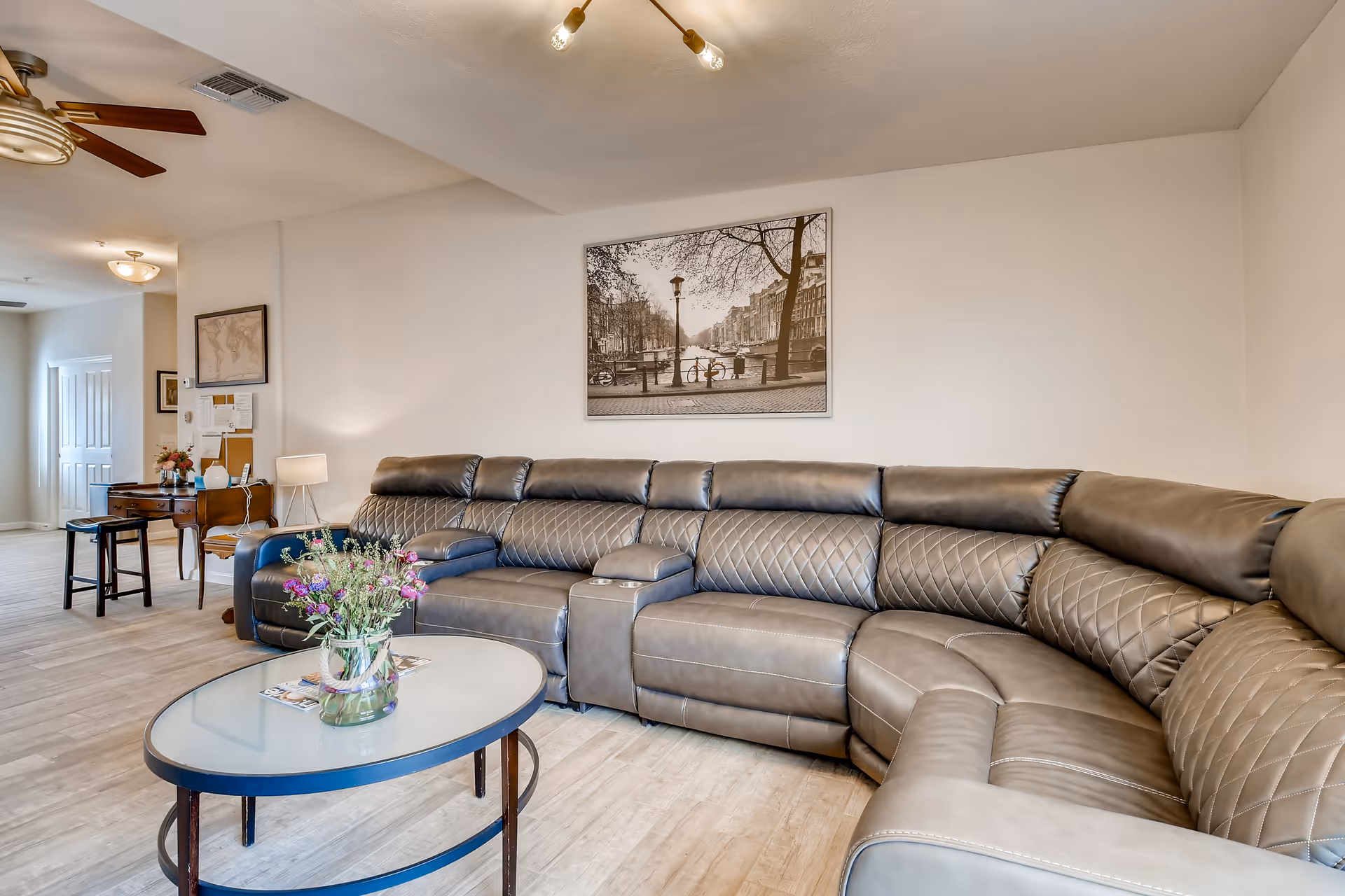 A spacious living room with a large, curved gray leather sectional sofa. In front of the sofa is an oval glass coffee table with a vase of flowers and some magazines. The walls are light-colored, and a black and white framed photograph of a canal scene hangs above the sofa. In the background, there is a small desk with a chair, a lamp, and some decorations, along with a ceiling fan and light fixtures.