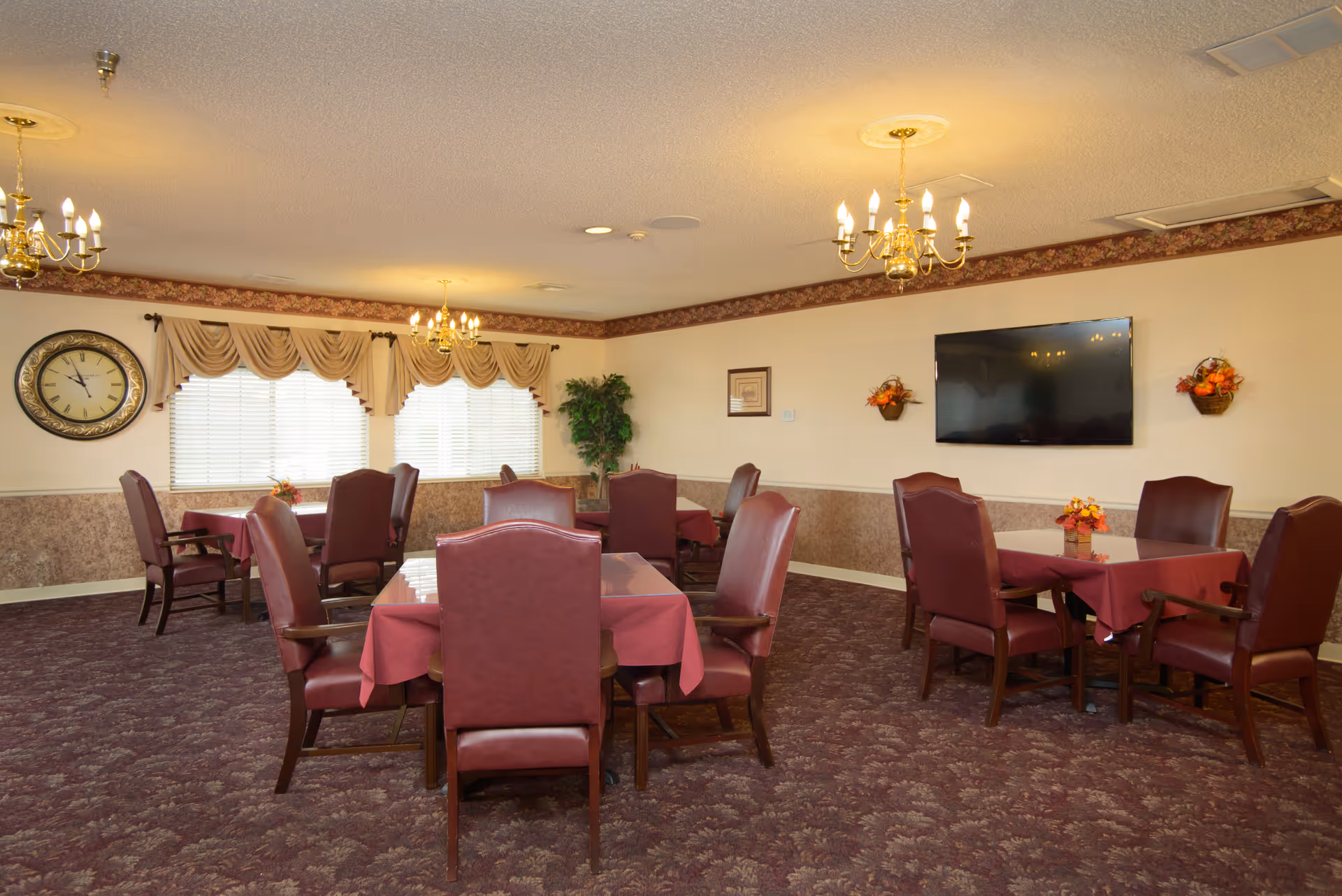 A dining room with several tables covered with red tablecloths, each surrounded by four maroon upholstered chairs. The room has patterned carpet, beige walls with a floral border near the ceiling, and three chandeliers hanging from the ceiling. There is a large clock on the wall near two windows with beige curtains, a flat-screen TV mounted on the wall, and two small wall-mounted flower arrangements.