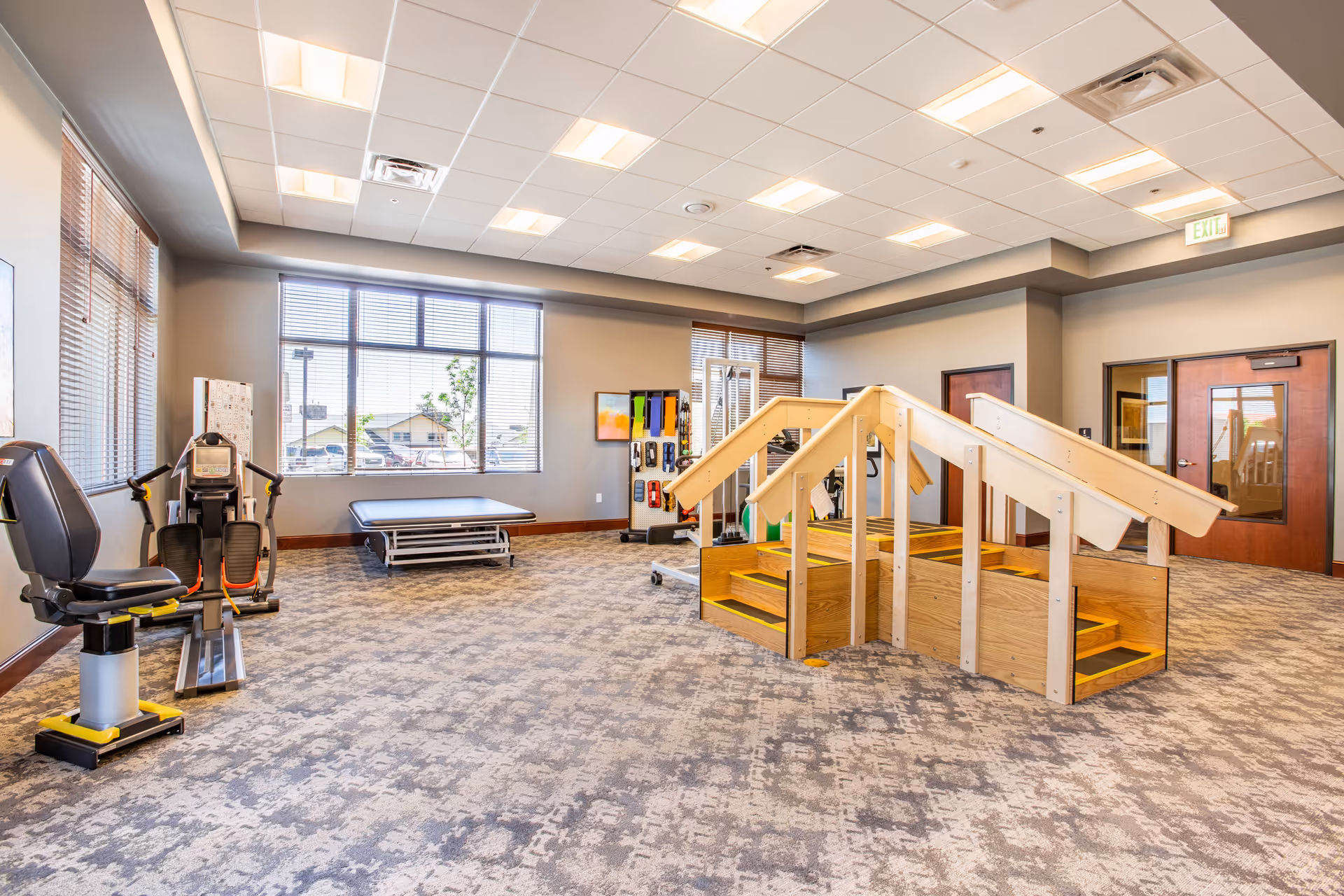 A spacious rehabilitation room with exercise equipment including a seated leg press machine, a padded therapy table, and a wooden staircase with handrails for physical therapy. Large windows allow natural light to fill the room, which has a patterned carpet and neutral-colored walls.