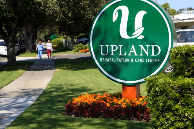 A large green circular sign with the text 'UPLAND REHABILITATION & CARE CENTER' stands on a well-maintained lawn with colorful flowers at its base. In the background, two people walk along a sidewalk lined with trees and parked cars.