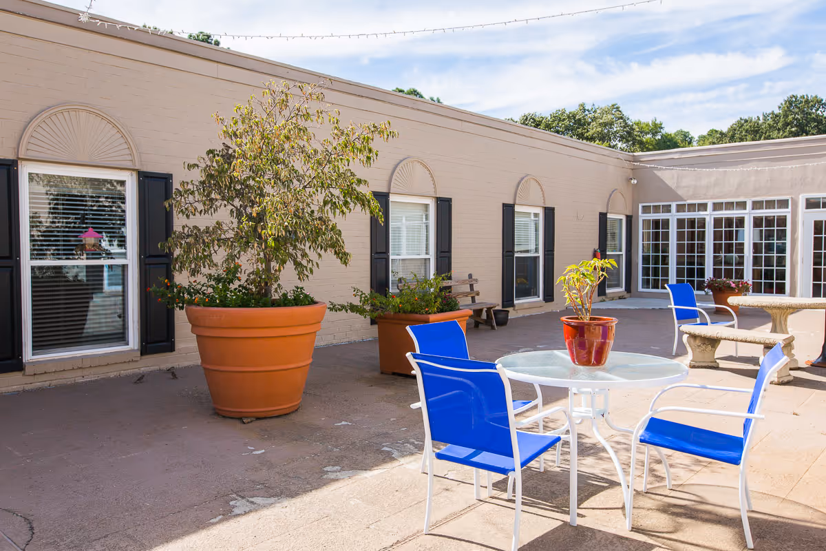 Outdoor patio area at Mallard Bay Nursing and Rehab with blue chairs around a glass table, large potted plants, and a beige building with windows and black shutters in the background under a partly cloudy sky.
