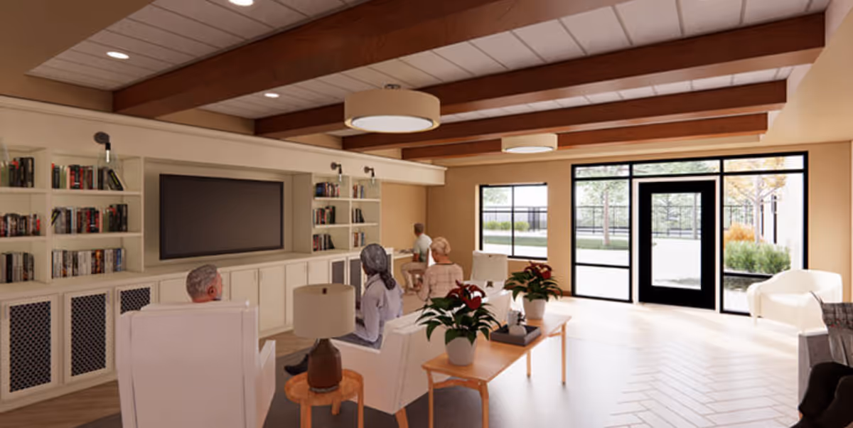 A bright community lounge with armchairs and sofas, a wall-mounted TV and bookshelves, and several residents seated facing the screen.