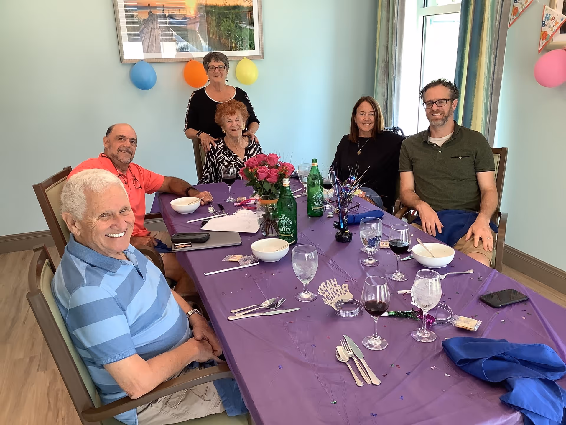 Six people gathered and smiling around a purple-covered dining table set with glasses, bowls, flowers, and party decorations in a light blue room.