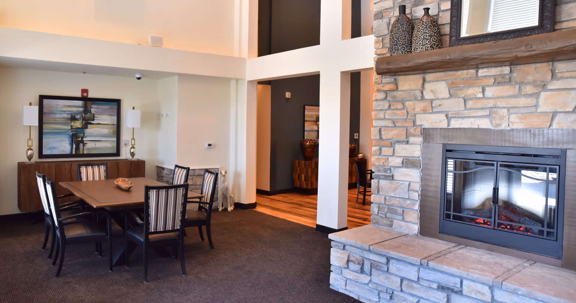 A cozy interior common area featuring a stone fireplace with decorative vases on the mantel. To the left, there is a dining table with six chairs, a sideboard with two lamps, and a large abstract painting on the wall. The space has a warm and inviting atmosphere with carpeted flooring and an open layout.