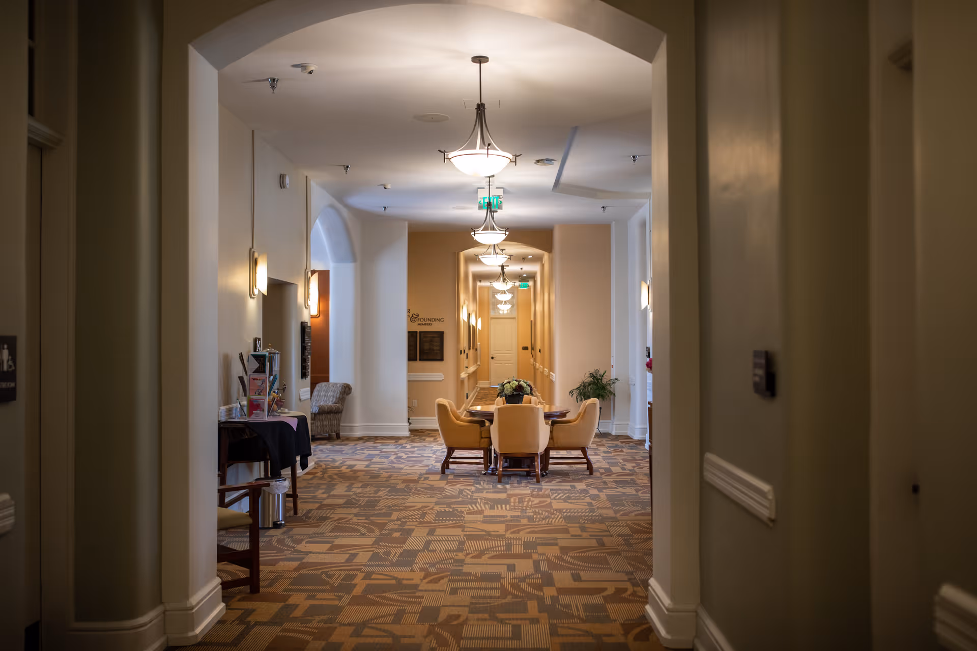 View down a well-lit hallway with patterned carpet in a senior living facility. The hallway features a round table with four cushioned chairs in the center, a few plants, wall sconces, and ceiling lights. There is an archway framing the view and a sign on the far wall.
