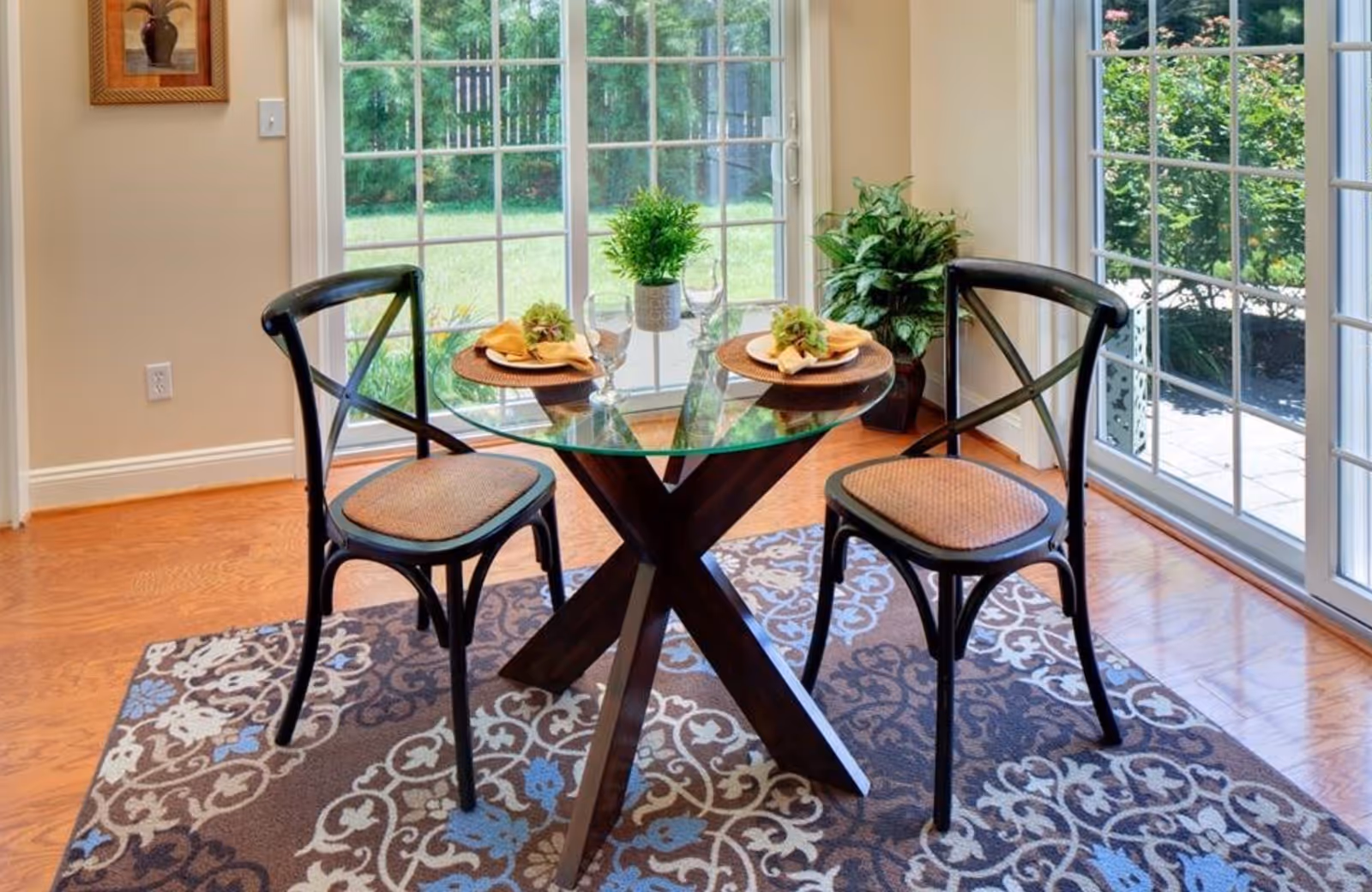 A small dining area with a round glass table set for two, featuring two black chairs with woven seats. The table has two place settings with plates, napkins, and glasses, along with a small green plant centerpiece. The area is well-lit by large sliding glass doors that open to a green outdoor space. A patterned rug is underneath the table and chairs, and there is a potted plant in the corner near the door.