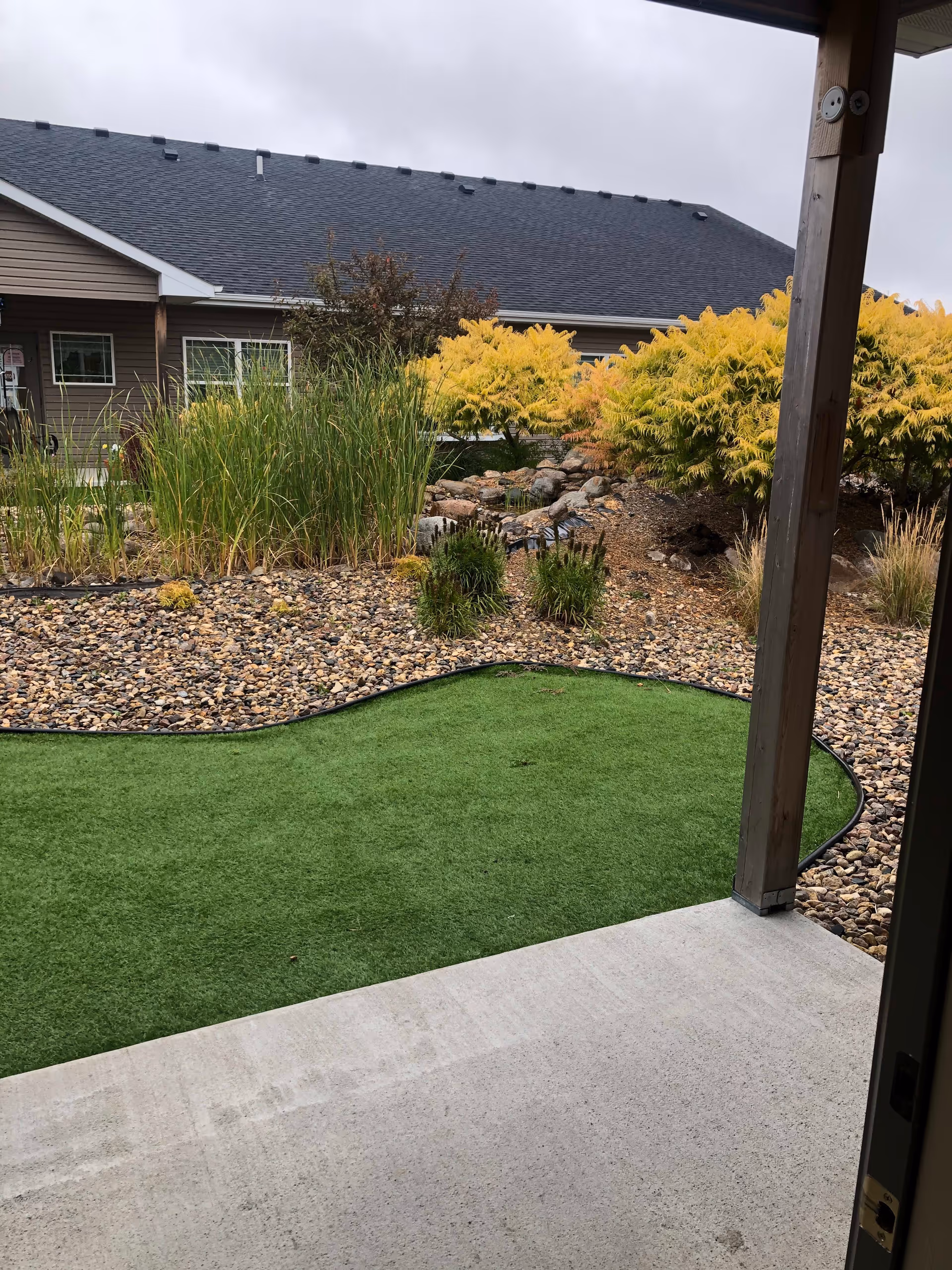 View from a covered patio looking out onto a landscaped garden area with green artificial turf, rocks, various plants, and shrubs. A building with a dark roof and beige siding is visible in the background under an overcast sky.