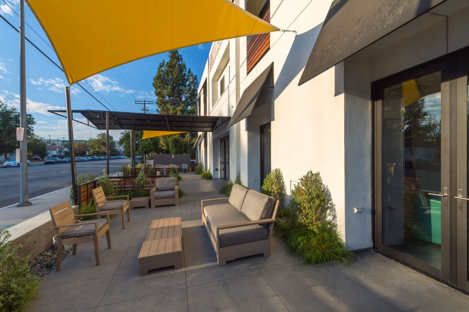 Outdoor patio area at AvantGarde Senior Living with modern wooden chairs, a cushioned sofa, and tables under yellow and black shade sails. The patio is adjacent to a building with large glass doors and small plants along the wall. A street with parked cars and trees is visible in the background under a partly cloudy sky.