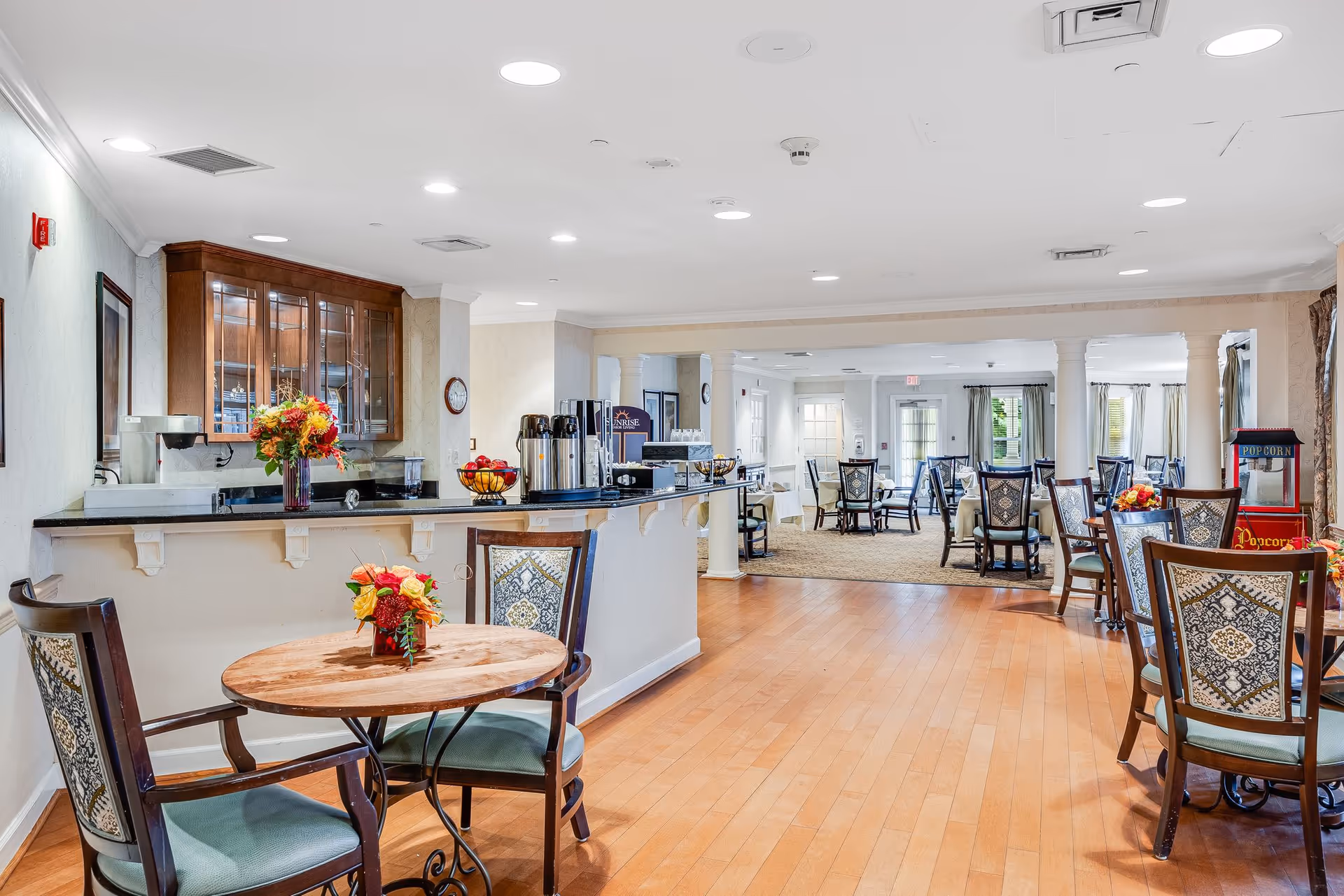 A spacious dining area in a senior living facility with wooden floors, multiple tables and chairs, a counter with coffee dispensers, a popcorn machine, and floral centerpieces on the tables. The room is well-lit with recessed ceiling lights and has large windows with curtains.