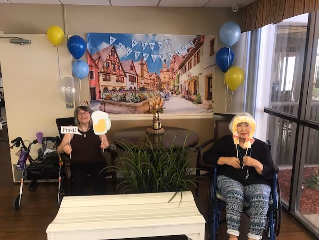 Two elderly women sitting indoors in a decorated room. One woman is holding a sign that says 'Prost!' and a cutout of a beer mug, while the other woman is holding a cutout of a person wearing a traditional hat. There are blue and yellow balloons on either side of a wall mural depicting a quaint European village scene. A white bench and a green plant are in the foreground.