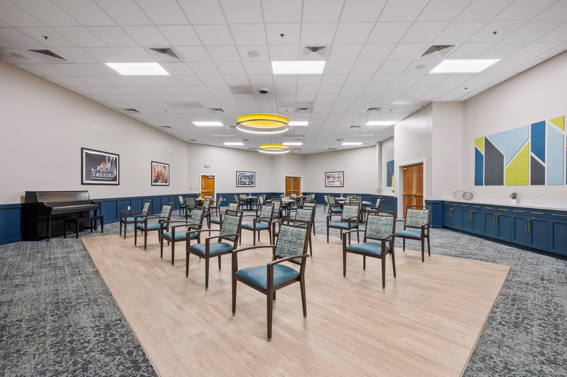 A spacious room with multiple chairs arranged in rows on a wooden floor section, surrounded by carpeted flooring. The walls are light-colored with blue wainscoting and decorated with framed pictures and colorful geometric wall art. There is a black piano with a bench on the left side and two large circular yellow ceiling lights. Several wooden doors are visible along the walls.