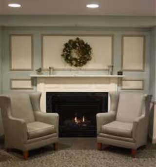 Two beige armchairs facing each other in front of a lit fireplace with a decorative wreath hanging above it on a light-colored wall.