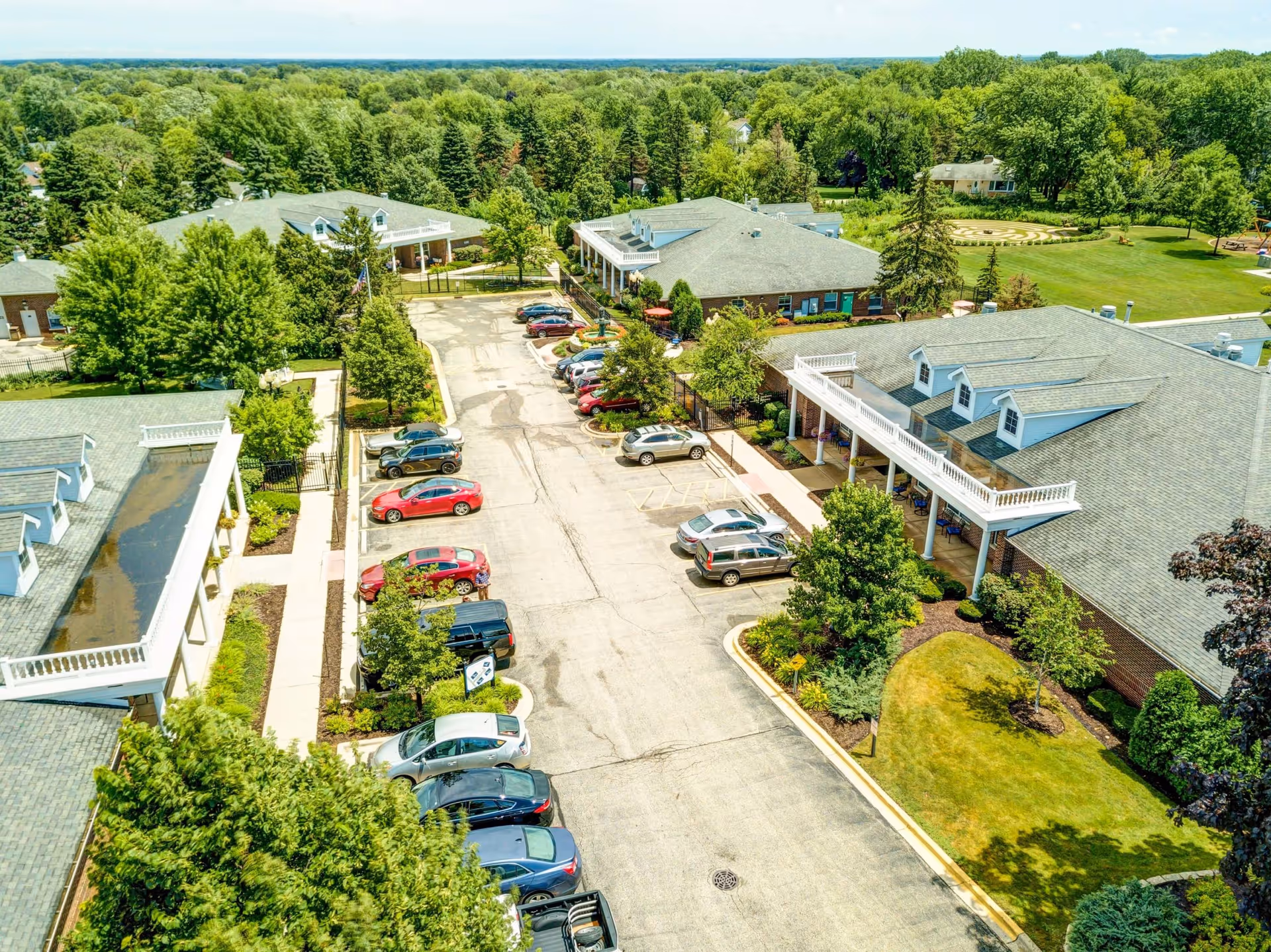 Aerial view of The Auberge at Lake Zurich senior living facility showing multiple buildings with gray roofs, a parking lot with several parked cars, well-maintained green lawns, trees, and a garden area with a circular hedge design in the background.