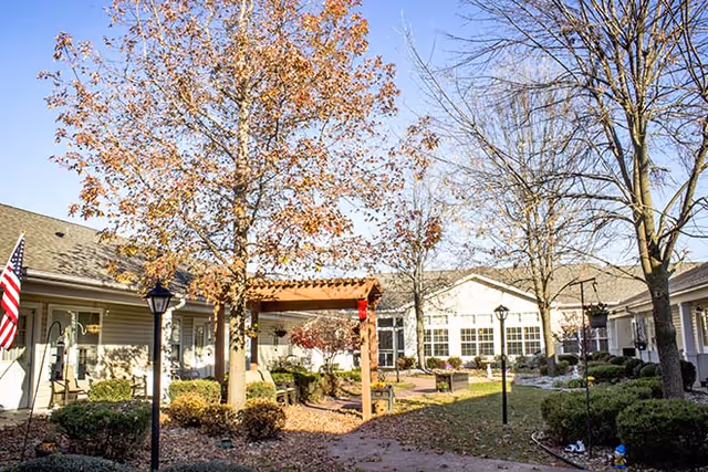 Sunny courtyard with a paved walkway, trees, a pergola and surrounding single-story senior living buildings.