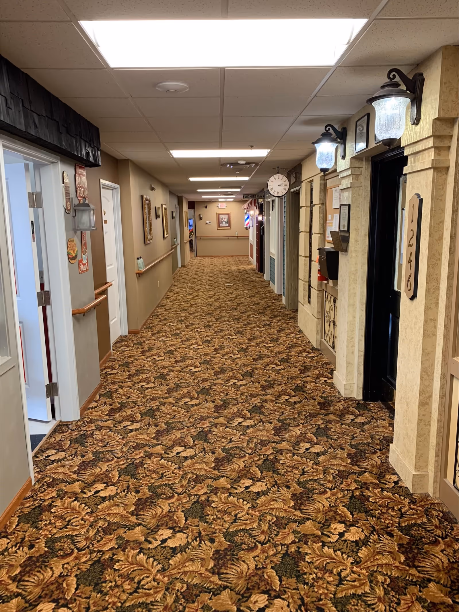 A long indoor hallway in a senior care facility with patterned carpet, beige walls, and ceiling lights. Doors line both sides of the hallway, some with room numbers and decorative elements. Handrails run along the walls, and a clock is mounted on the right side. The hallway appears clean and well-lit.