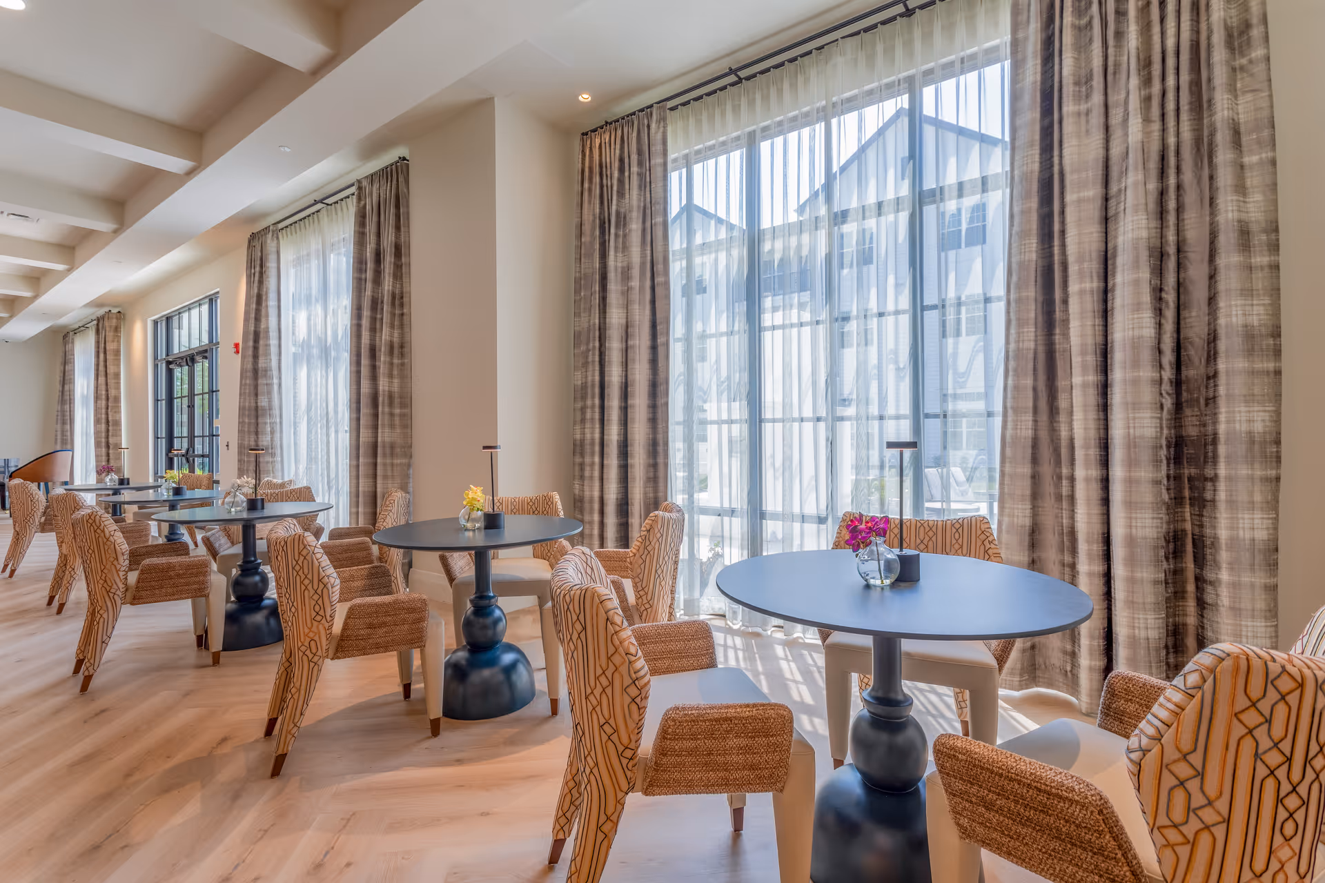 Bright dining area with round tables and patterned upholstered chairs arranged neatly. Large windows with sheer and heavy curtains allow natural light to fill the room. Small flower vases are placed on each table.