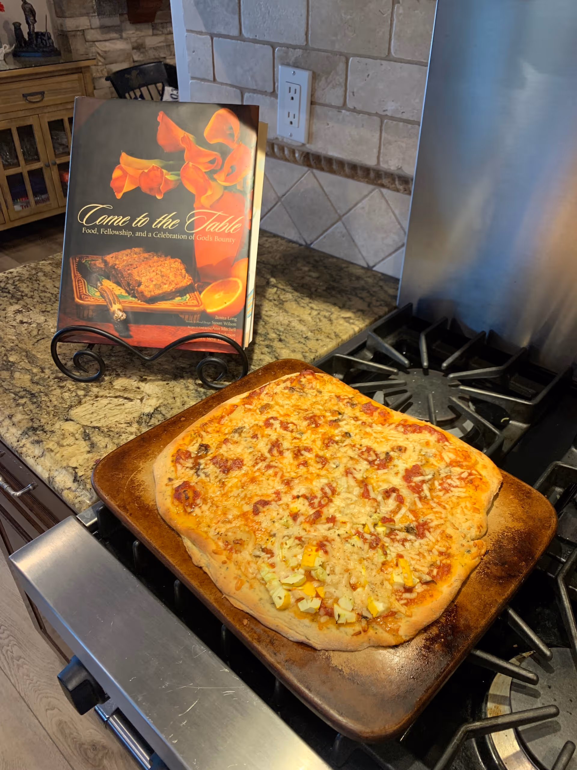 A freshly baked rectangular pizza on a baking tray placed on a gas stove in a kitchen. Behind the pizza, there is a cookbook titled 'Come to the Table: Food, Fellowship, and a Celebration of God's Bounty' displayed on a stand. The kitchen has granite countertops and a tiled backsplash.
