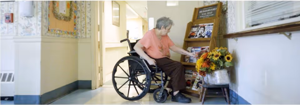 An elderly person in a wheelchair reaching for a magazine from a wooden rack in a hallway of a nursing facility. There is a small table with a flower arrangement next to the magazine rack, and the hallway has light-colored walls with a window on the right side.