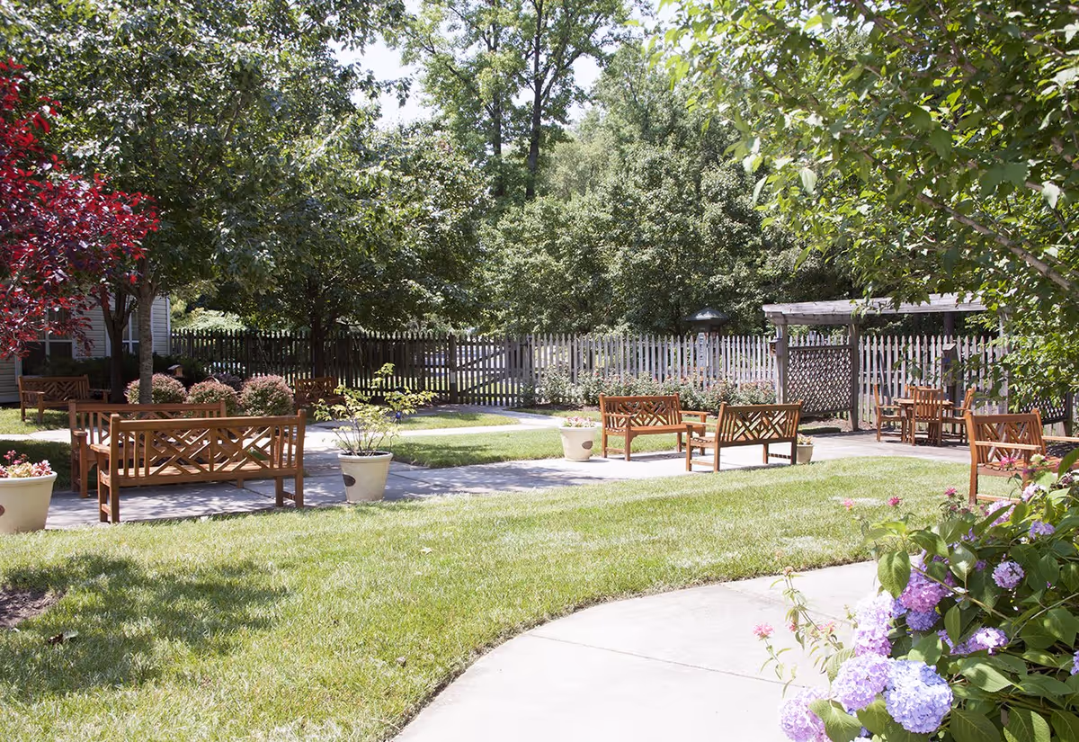 Sunny landscaped courtyard with wooden benches, potted plants, a pergola and a curved walkway surrounded by trees and flowers.