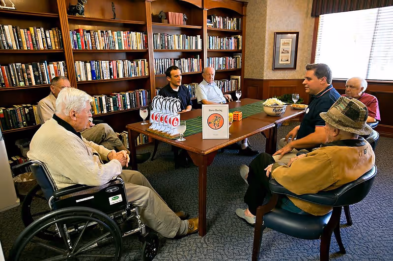 A group of elderly men and a younger man sitting around a rectangular table in a room with bookshelves filled with books. They appear to be engaged in a game or activity involving cards and dice. One elderly man is in a wheelchair, and the room is well-lit with a window covered by blinds.