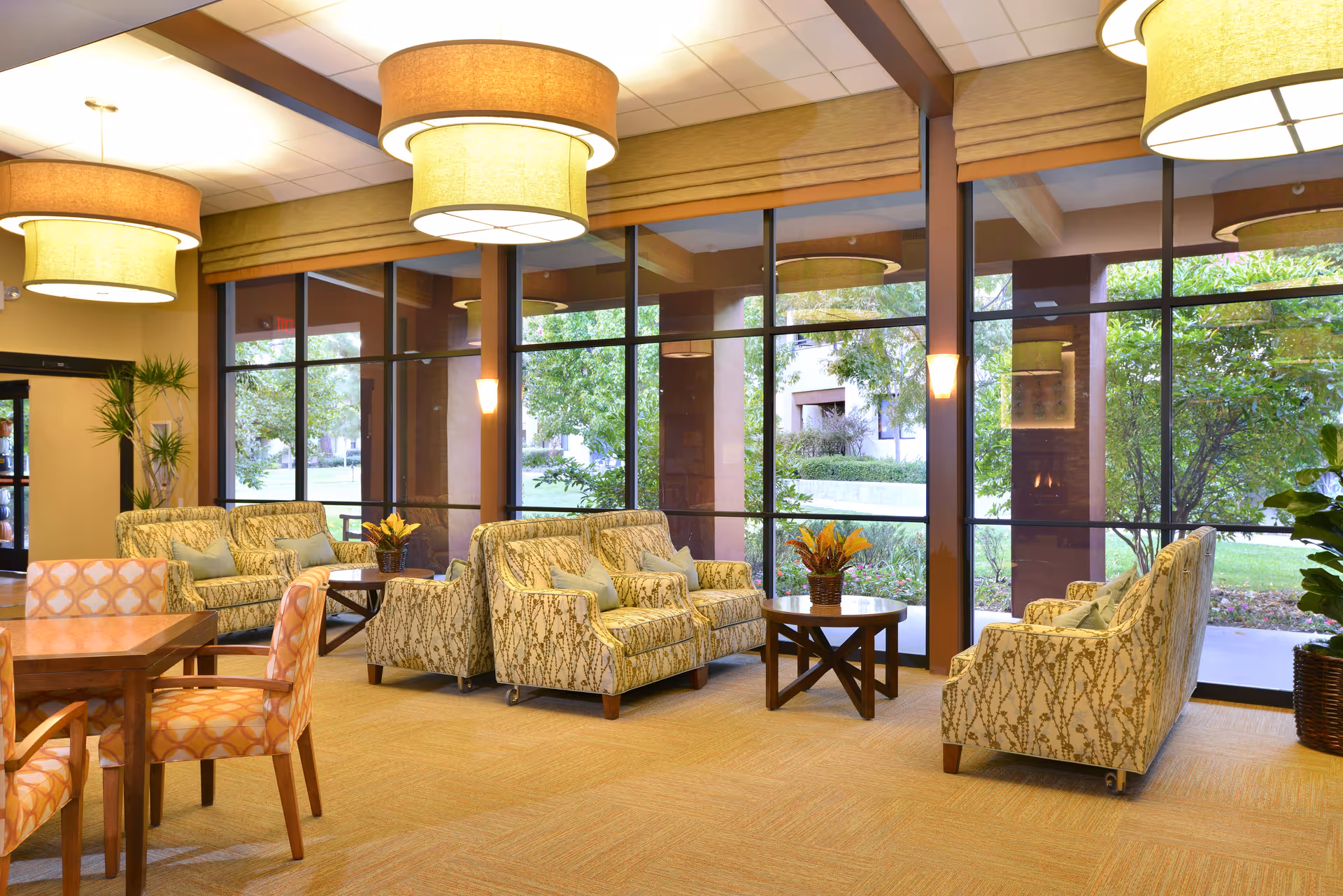 A bright and spacious living room area with large floor-to-ceiling windows showing a green outdoor garden. The room features patterned sofas and armchairs arranged around wooden coffee tables with flower arrangements. There are also dining tables with patterned chairs, large round ceiling lights, and potted plants near the entrance.