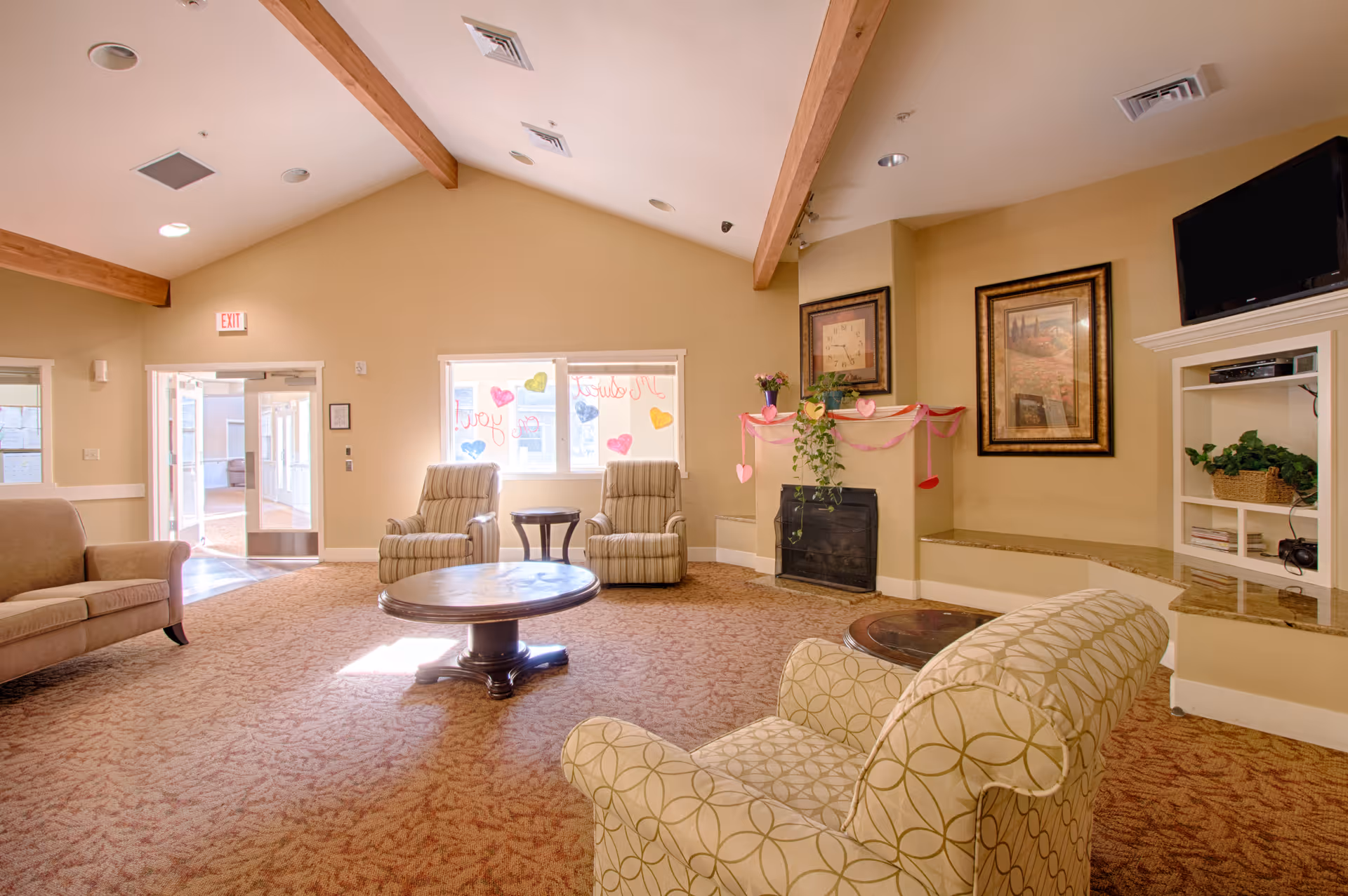 A cozy living room in a memory care community with beige walls and carpet. The room features a patterned armchair in the foreground, two striped armchairs near a window decorated with colorful heart-shaped drawings, a round wooden coffee table, a fireplace adorned with pink and red paper hearts and streamers, framed artwork on the walls, and a mounted flat-screen TV above a built-in shelving unit with plants and books.