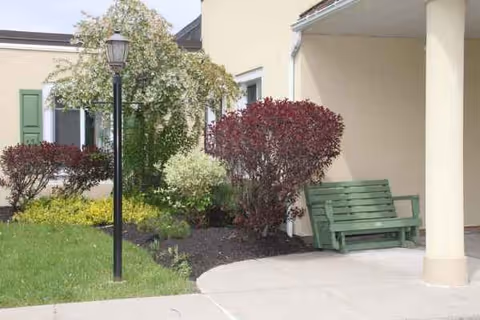 A green bench sits beneath a covered area beside landscaped shrubs and a lamppost.