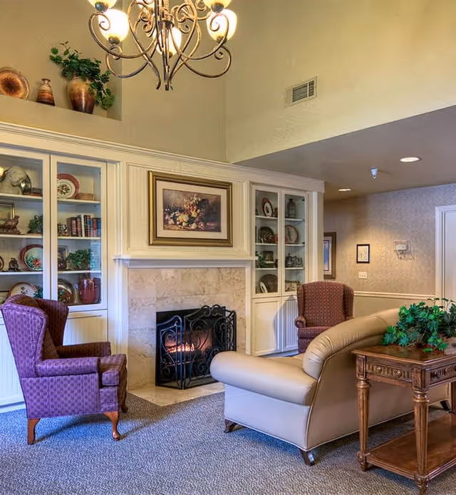 A cozy living room area with a lit fireplace surrounded by built-in white shelves displaying decorative plates, books, and plants. The room features a beige leather sofa, two purple upholstered armchairs, a wooden side table with a green plant, and a chandelier hanging from the ceiling.