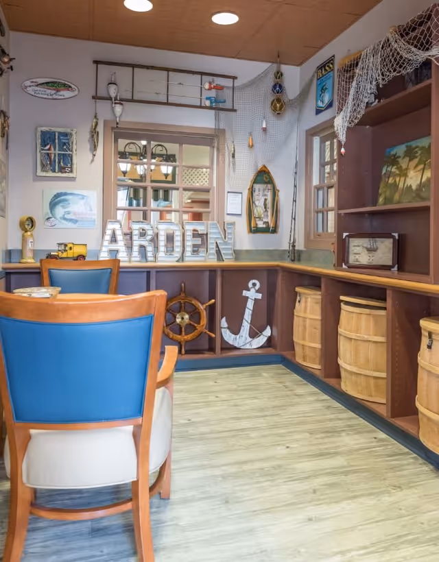 Interior room with nautical-themed decor including a ship's wheel, anchor, fishing nets, and barrels. There are two wooden chairs with blue cushions facing a counter with the letters 'ARDEN' displayed. The walls have various fishing-related decorations and a window showing another room.