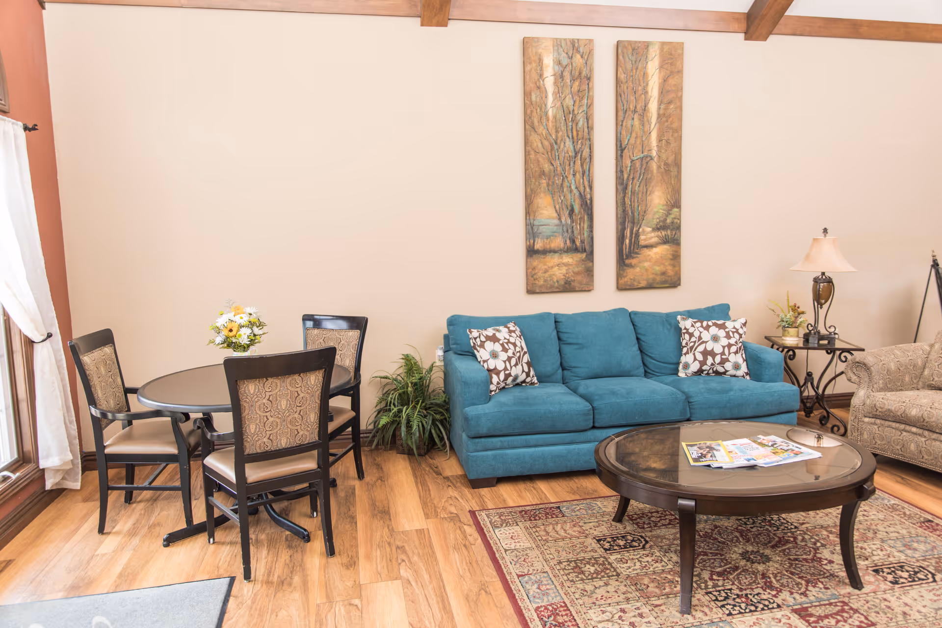 A cozy living room area with a teal sofa adorned with two floral patterned pillows, a round glass coffee table with magazines on top, and a patterned armchair. To the left, there is a small round dining table with four chairs and a vase of flowers. The room has wooden flooring, a decorative rug, two vertical paintings of trees on the wall, and a side table with a lamp and a small plant.
