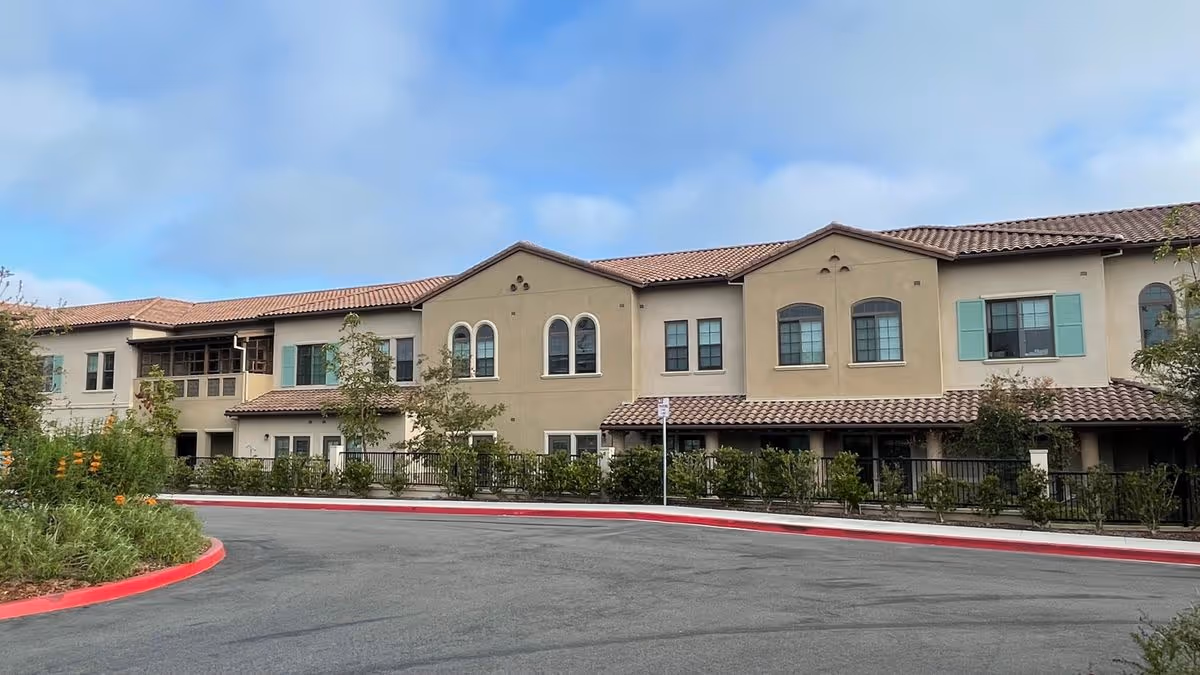 Two-story beige Mediterranean-style building with a red-tiled roof facing a curved driveway and landscaped greenery.