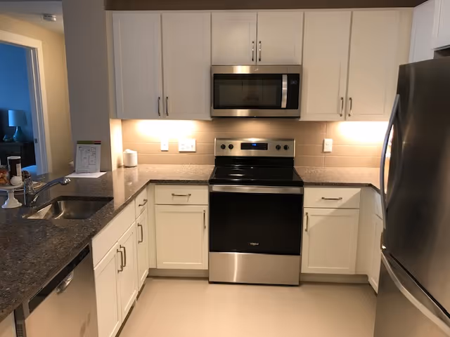 Modern kitchen with white cabinets, stainless steel appliances (range, microwave, refrigerator), gray countertops and under-cabinet lighting.