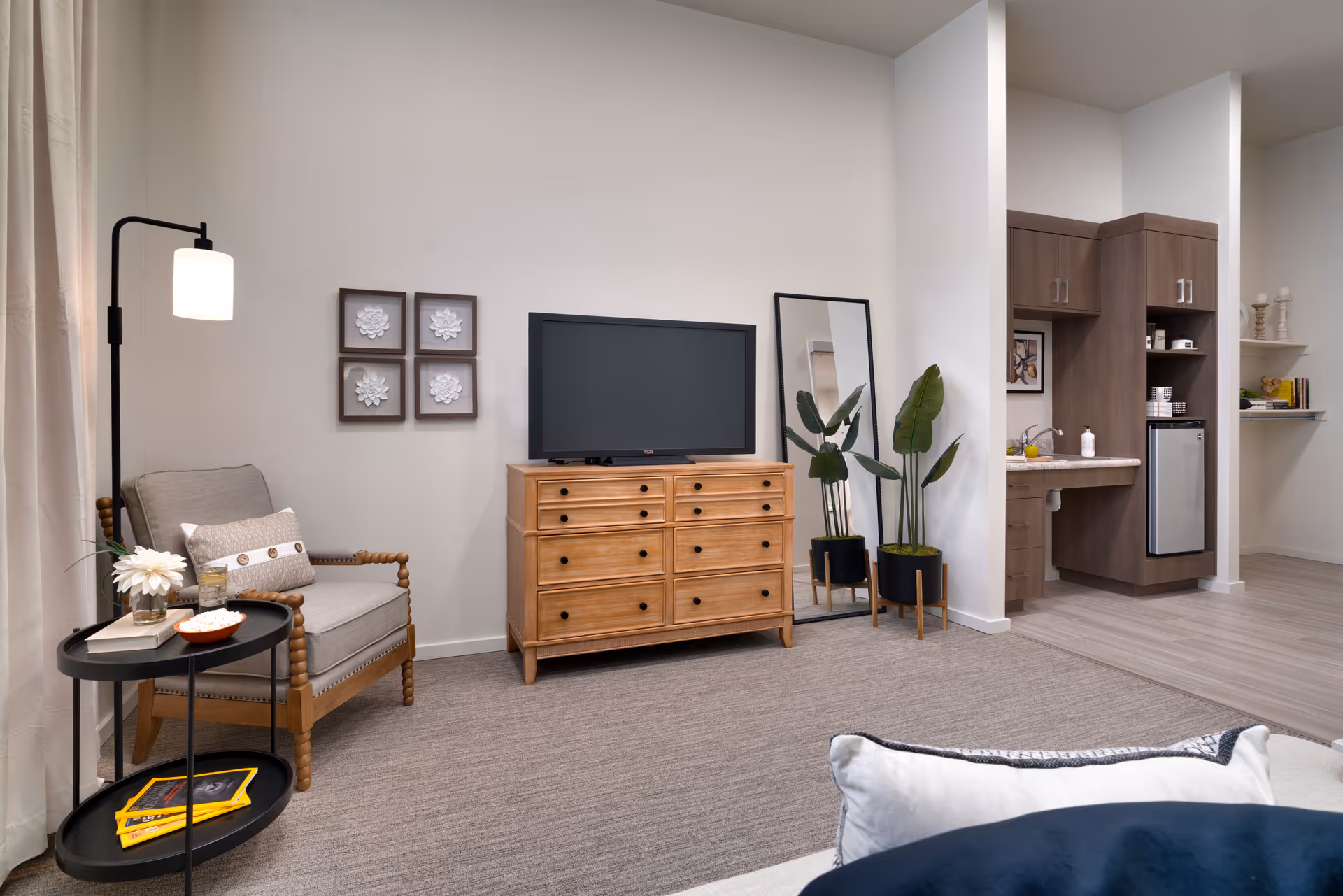 A cozy living area in a senior living facility featuring a wooden armchair with cushions next to a black floor lamp and a round black side table with books and a flower vase. A wooden dresser with a flat-screen TV on top is against the wall, accompanied by a tall mirror and two potted plants. In the background, a small kitchenette with wooden cabinets, a sink, and a mini fridge is visible.