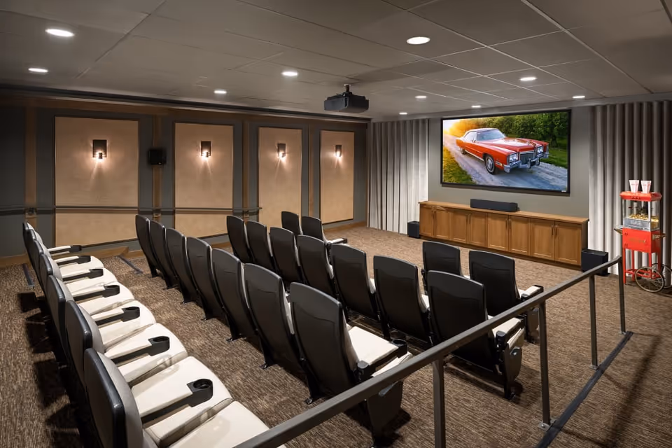 A small theater room with three rows of black and white cushioned seats facing a large screen displaying a red classic car. The room has beige walls with decorative panels, wall sconces, a popcorn machine on the right side, and a wooden cabinet beneath the screen.