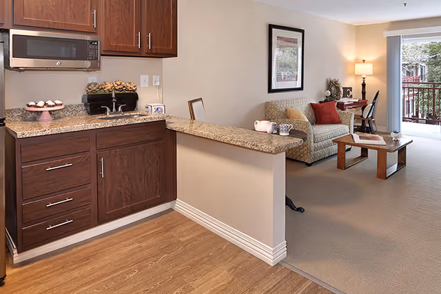 Interior view of a senior living facility apartment showing a small kitchen area with dark wood cabinets, a microwave, and granite countertops. Adjacent to the kitchen is a living room with a patterned sofa, red and beige cushions, a wooden coffee table, a desk with a chair and lamp, and a sliding glass door leading to a balcony.