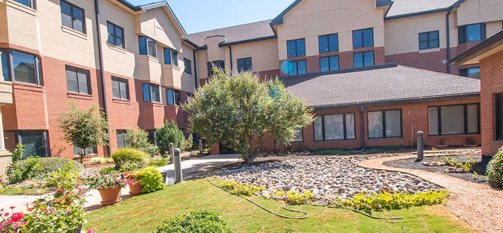 Landscaped courtyard with a rock garden, potted flowers, shrubs and surrounding multi-story brick senior living building.