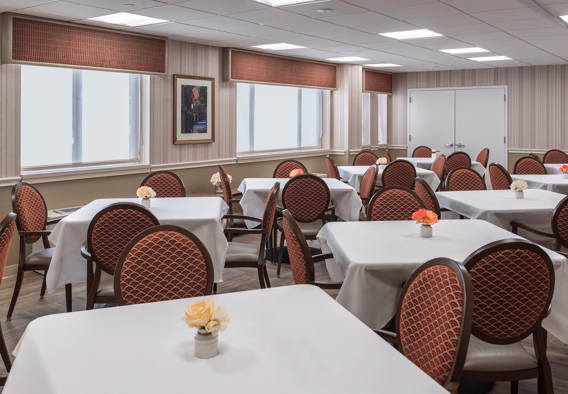 A bright dining room with multiple round-backed chairs and tables covered in white tablecloths, each topped with small flower arrangements.