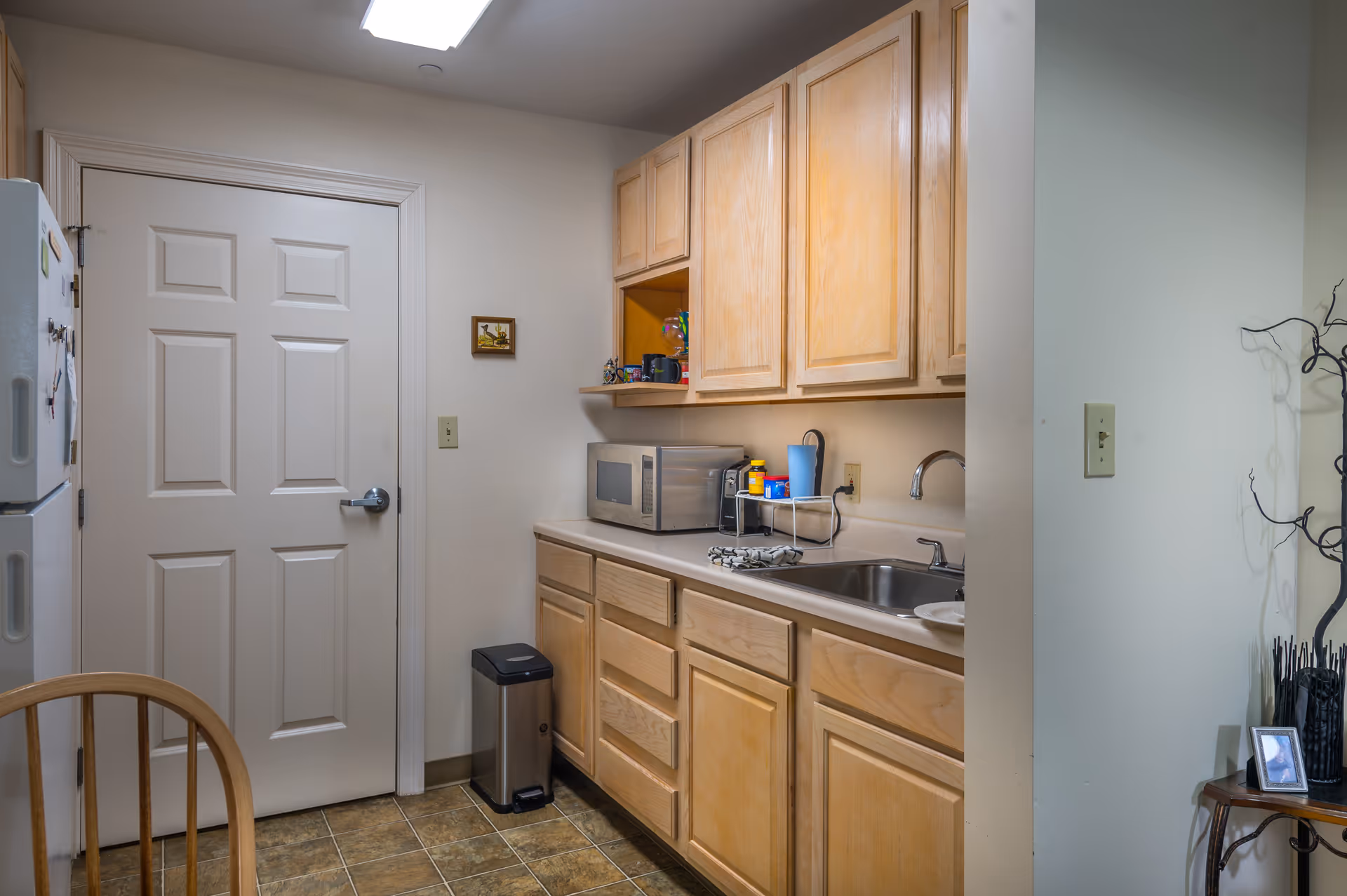 Compact kitchen area with light wood cabinets, countertop microwave and sink, a refrigerator, and a small trash can.