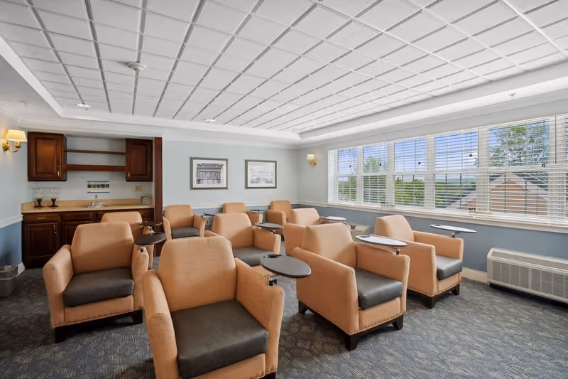 A bright room with multiple beige armchairs arranged in rows, each equipped with a small attached table. The room has large windows with white blinds letting in natural light, a patterned carpet, and a small kitchenette area with wooden cabinets and a sink in the corner.